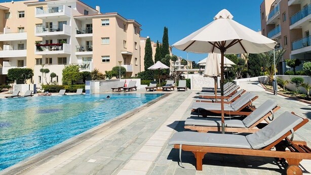 A swimming pool with lounge chairs and umbrellas, surrounded by multi-story buildings with balconies. - Luxury 2BR Apt at Elysia Park S6 with Pools, Gym & Spa - Center Paphos (Pafos)