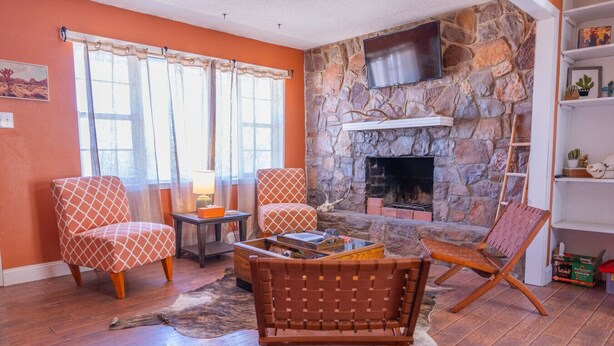 Uma sala de estar com lareira de pedra, dois poltronas estampadas, uma mesa de centro e uma estante.