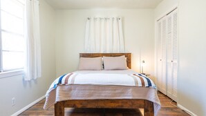 Quarto com cama de madeira, cortinas brancas, abajur e armário.