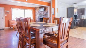Uma sala de jantar com mesa e cadeiras de madeira, lareira de pedra e cozinha ao fundo.