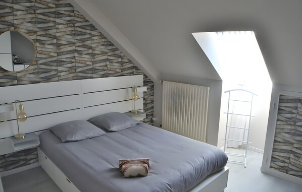 Een slaapkamer met een schuine kap, een bed met grijze beddengoed, een nachtkastje met een lamp, een spiegel en een raam met uitzicht.