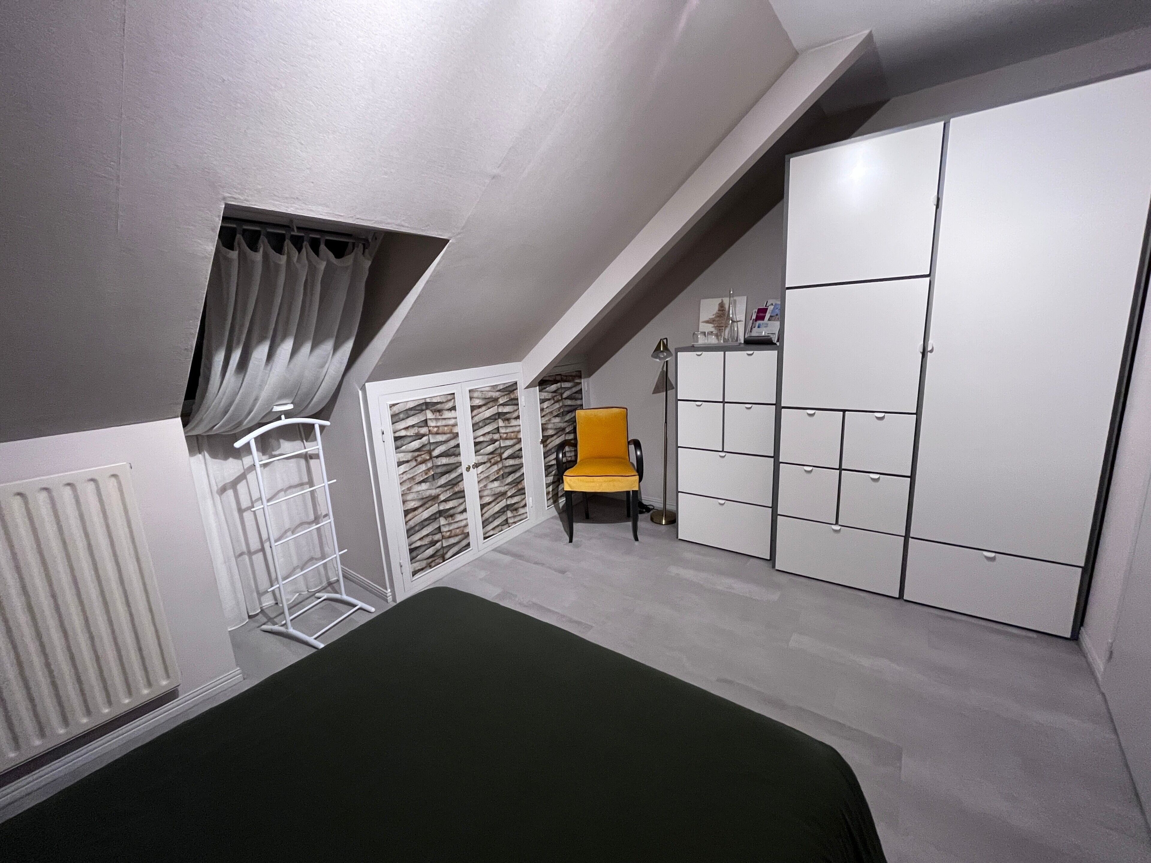 A bedroom with a sloped ceiling, a bed, a chair, a wardrobe, and a radiator.