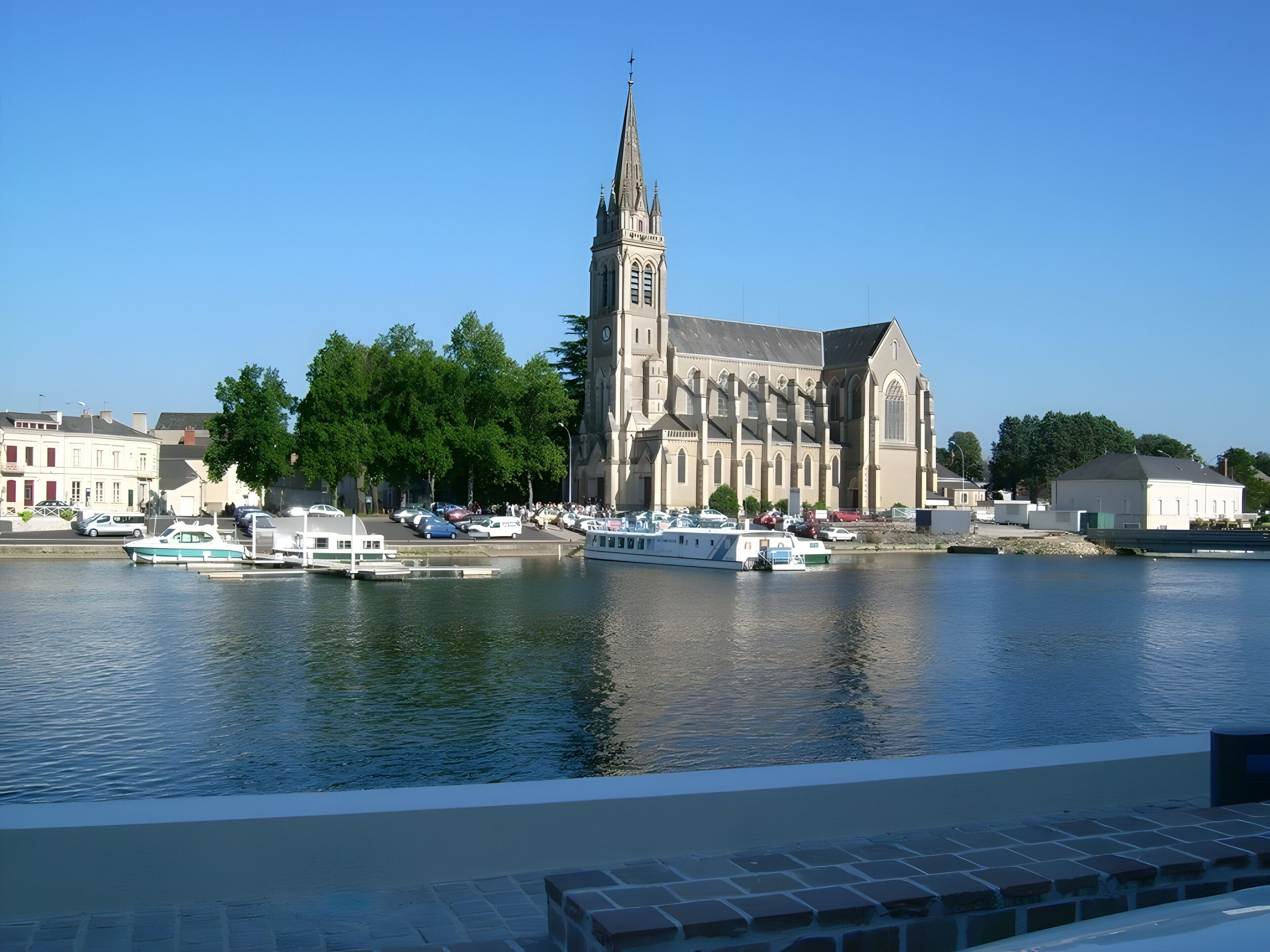 Une église historique dotée d’un clocher imposant, une rivière où naviguent des bateaux et, au premier plan, un pont de pierre.