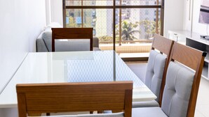 A modern dining area with a glass table and wooden chairs, large windows with a city view, and a flat-screen TV on a stand. - Buenos Ayres #504 - Comfortable Apartment by Carpediem (João Pessoa)