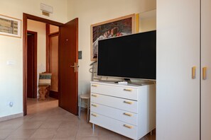 Une pièce avec un téléviseur posé sur un meuble blanc, une porte donnant sur une autre pièce et un petit tabouret.