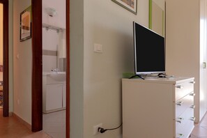 Un couloir avec un meuble blanc, un téléviseur posé sur le dessus, et une salle de bain avec un lavabo et un miroir.