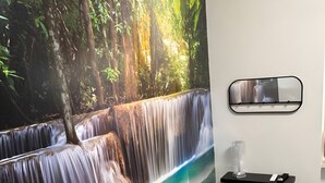 A living room with a large waterfall mural, a sofa, a black pillow, and a wall-mounted shelf with a glass vase and remote controls. - Holiday Home 'Genghetta' with Garden, Wi-Fi and Air Conditioning (Rivotorto)