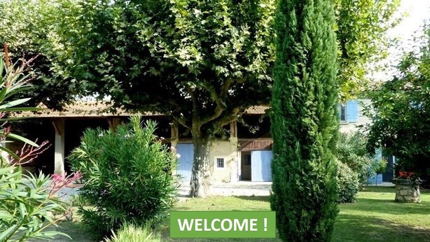 Un jardín con un letrero de bienvenida, un edificio de techo a dos aguas y diversa vegetación.