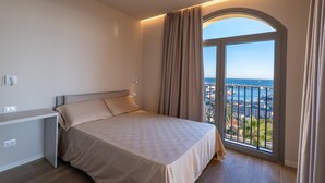 A bedroom with a bed, a desk, and a large window with a view of the sea and buildings. - Villa Pignone 12 (Genova)