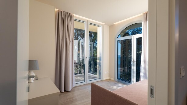 A modern room with a sliding glass door leading to a balcony, a small bedside table with a lamp, and a large arched window. - Villa Pignone 12 (Genova)