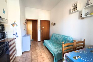 A compact kitchen with a microwave, stove, and a small refrigerator. There is a wooden door leading to another room, a blue sofa, and a table with a patterned tablecloth. - CV019 – Cervia, one-bedroom apartment with balcony and parking space (Cervia)