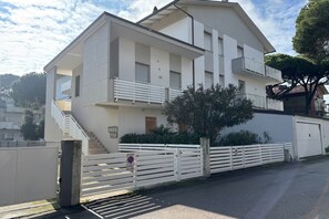 Un bâtiment moderne à deux étages, doté d’une façade blanche, d’un escalier menant à l’étage supérieur et d’une entrée fermée par un portail.
