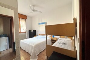 Une chambre d’hôtel avec deux lits, un ventilateur de plafond, un radiateur, une fenêtre avec des rideaux, un bureau avec une lampe et un mur orné d’un tableau.
