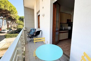 Un balcon avec une table et des chaises, une porte-fenêtre coulissante donnant sur une cuisine, et une vue sur les voitures garées et les arbres.