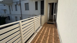 Ein Balkon mit weißer Brüstung, gefliestem Boden und einem Gebäude mit mehreren Fenstern und Balkonen im Hintergrund.