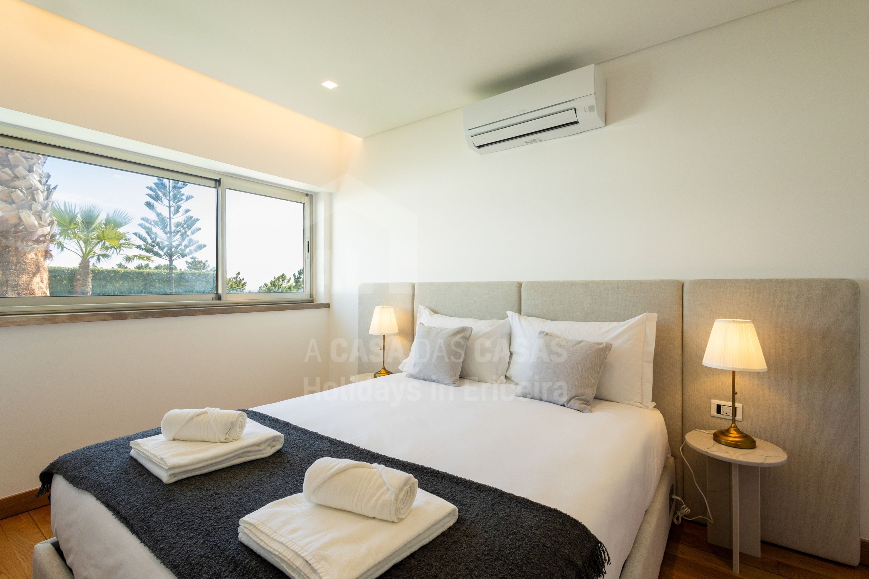Une chambre d’hôtel avec un grand lit, deux lampes de chevet, une fenêtre donnant sur des palmiers et un climatiseur fixé au mur.