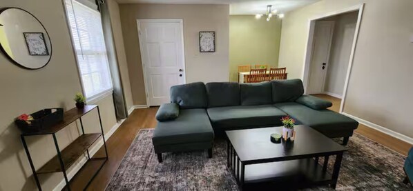 A living room with a sectional sofa, a coffee table, and a side table. There is a dining area visible in the background.