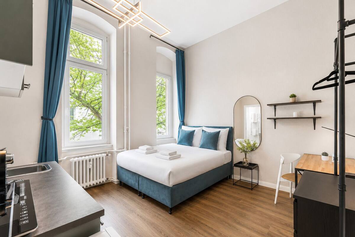 A modern hotel room with a bed, a desk, a chair, a radiator, and a window with curtains.