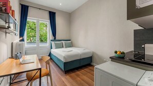 Habitació petita amb un llit, un escriptori amb un portàtil, una rentadora i una finestra amb cortines.