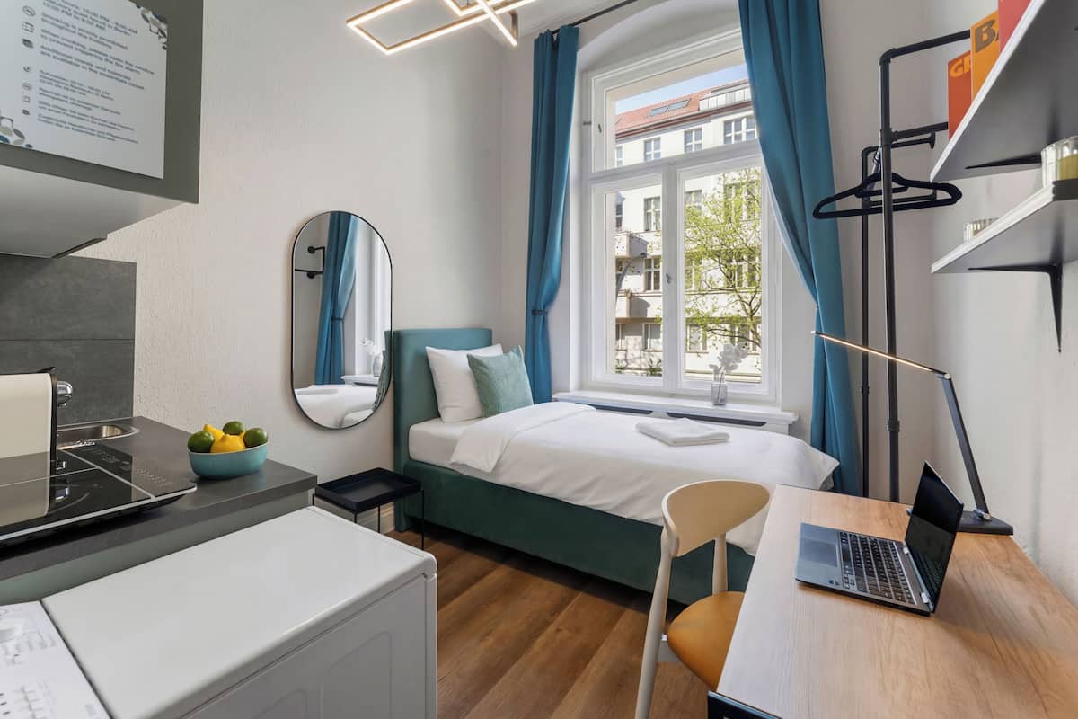 A modern hotel room with a bed, a desk with a laptop, a bowl of fruit, a mirror, and a window with curtains.