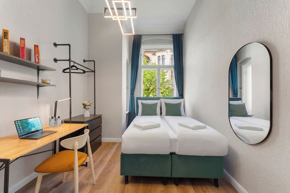 A modern hotel room with a bed, a desk with a laptop, a chair, a mirror, and a view of the outside.