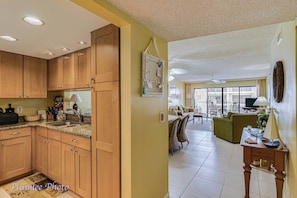A spacious living area with a dining table, chairs, and a sofa. A kitchen with wooden cabinets and a sink is visible on the left. - Golden Shores 516 - Intercoastal Views (Indian Shores)