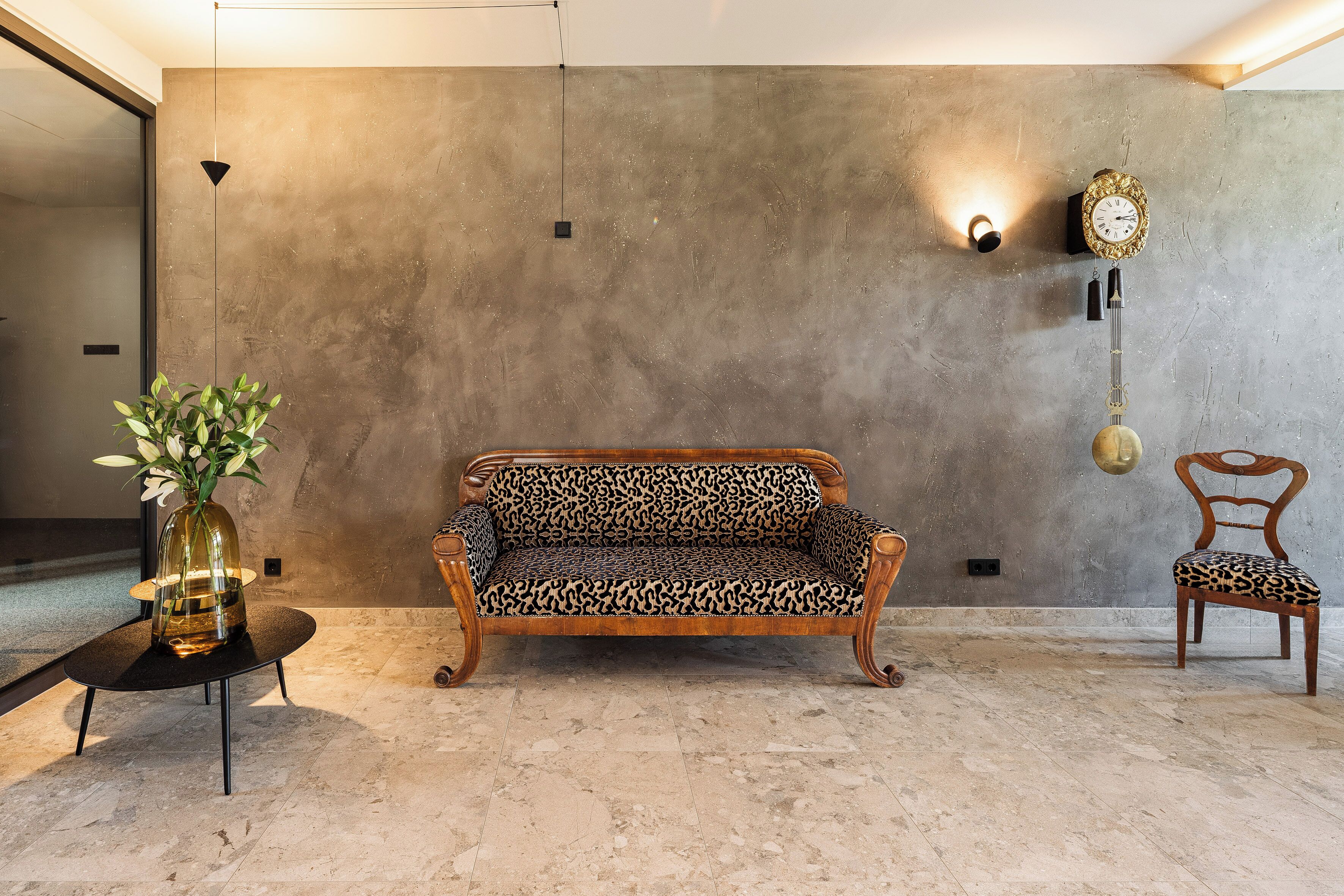 A room with a sofa, a chair, a wall-mounted clock, and a vase with flowers.