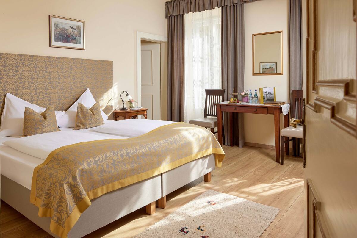 Une chambre d’hôtel avec un grand lit, un bureau et une chaise. Il y a une fenêtre avec des rideaux, un lustre et un tableau encadré au mur.
