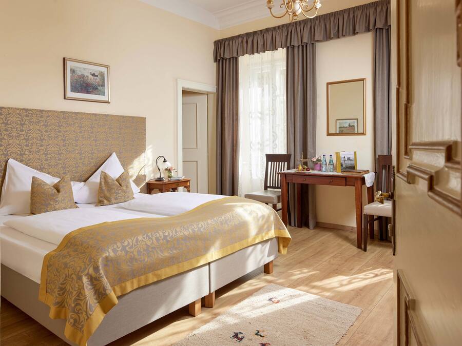 A hotel room with a large bed, a desk, and a chair. There is a window with curtains, a chandelier, and a framed picture on the wall.