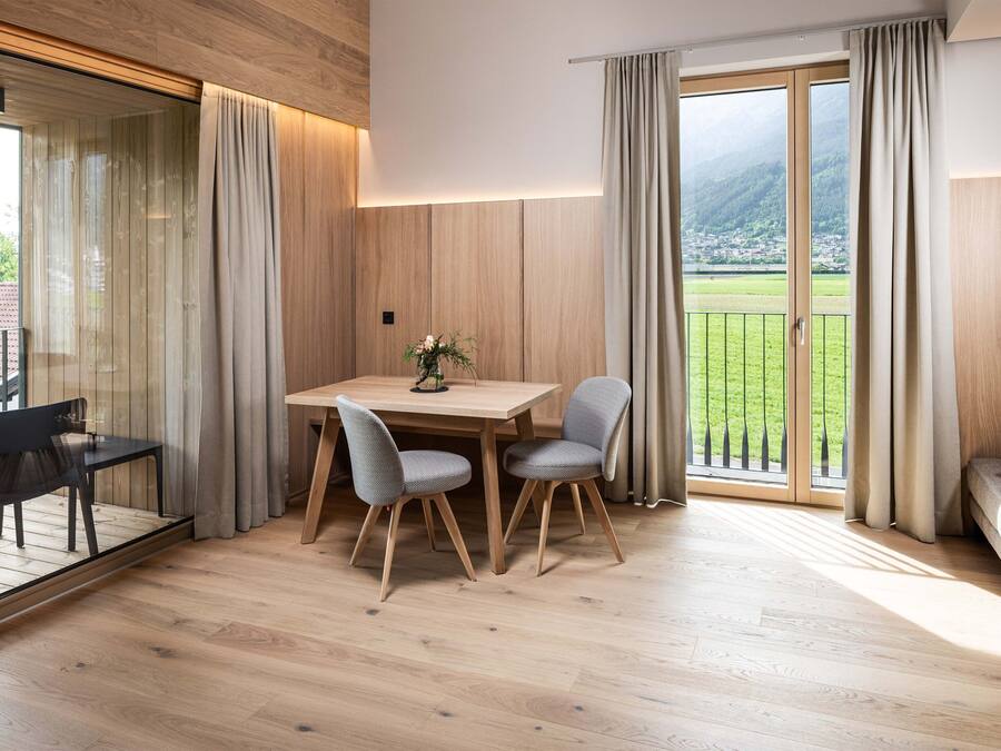 A modern dining area with a wooden table and chairs, a view of a green landscape through large windows, and a balcony with a railing.
