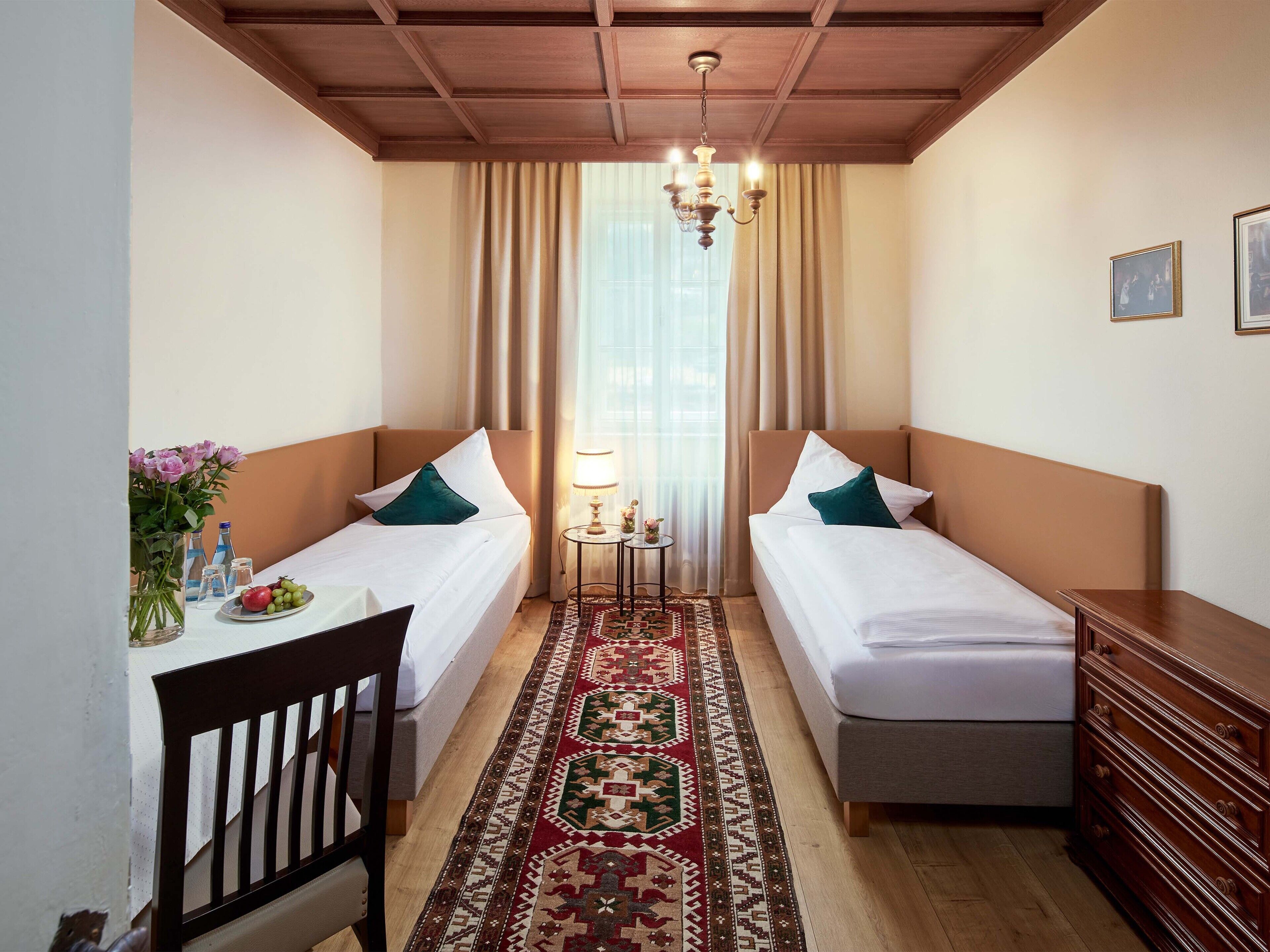 A hotel room with two beds, a wooden dresser, a small table with a lamp, a vase with flowers, and a patterned rug.