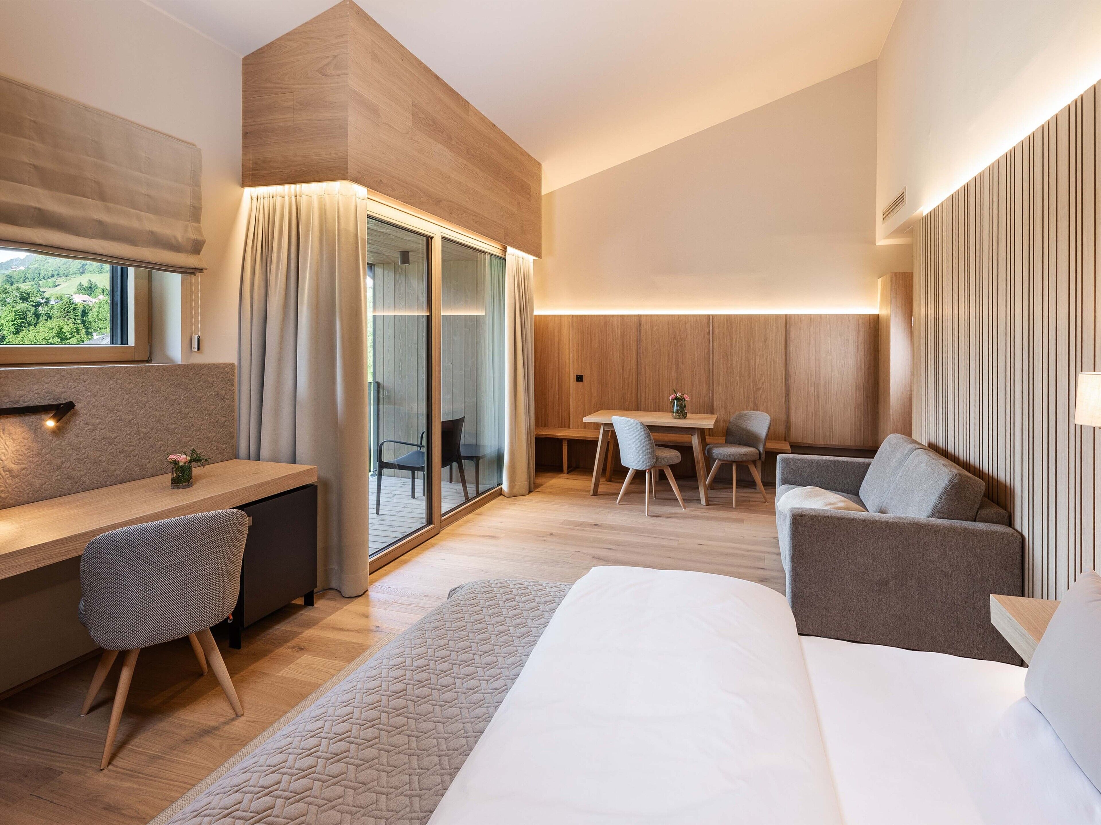 A modern hotel room with a wooden accent wall, a desk with a chair, a sofa, a dining table with chairs, and a large window with curtains.
