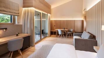 A modern hotel room with a wooden accent wall, a desk with a chair, a sofa, a dining table with chairs, and a large window with curtains.