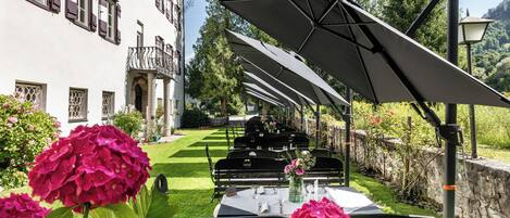 Un área de comedor al aire libre con mesas, sillas y sombrillas, rodeada de follaje y un edificio con ventanas.