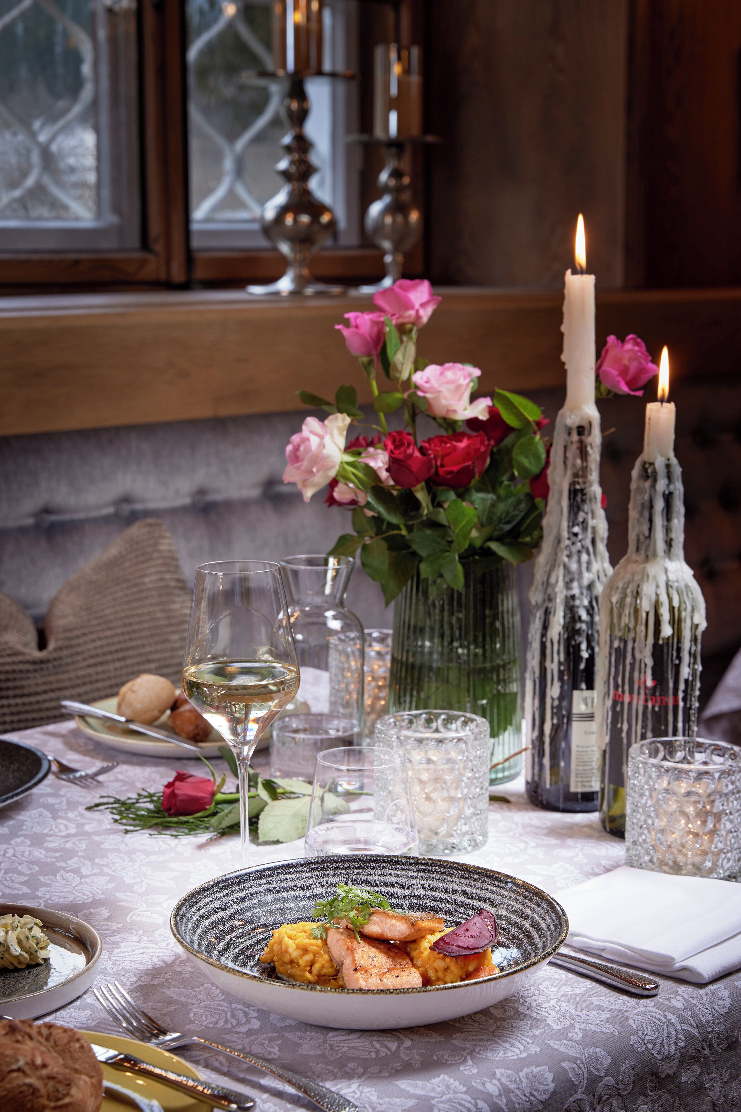 A dining table set with a plate of food, glasses, candles, and a vase of flowers.