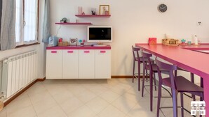 Eine kompakte Küche mit Essbereich, mit einem weißen Schrank mit pinken Griffen, einer weißen Arbeitsplatte und einem roten Esstisch mit violetten Stühlen.