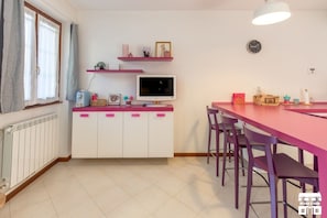 Cocina compacta con zona de comedor, que incluye un armario blanco con detalles rosa, una encimera blanca y una mesa de comedor roja con sillas moradas.