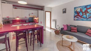 Eine moderne Küche mit Kochinsel, weißen Schränken und rosa Akzenten. Es gibt einen Essbereich mit Tisch und Stühlen, eine Sitzecke und ein Bild an der Wand.