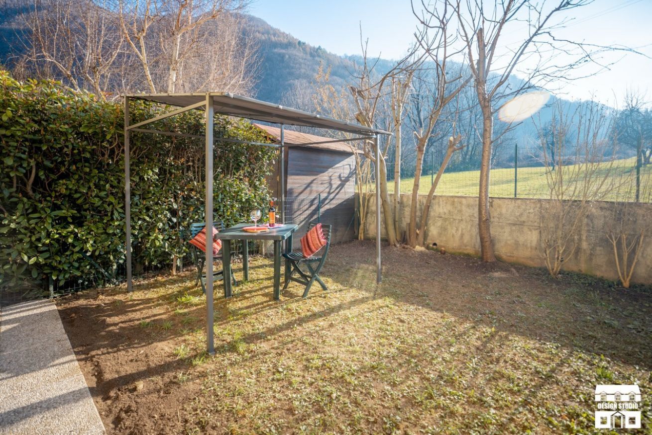 Zona salotto esterna riparata con tavolo e sedie, immersa nel verde e con vista sulle montagne.