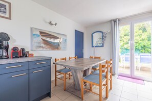 Un coin repas avec une table et des chaises en bois, une cuisine équipée de placards et d’appareils électroménagers, un tableau et une porte donnant sur un espace extérieur.