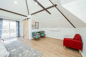 A spacious loft bedroom with a sloped ceiling, wooden beams, a large sliding glass door leading to a balcony, two floral armchairs, a red sofa, and a framed picture on the wall.