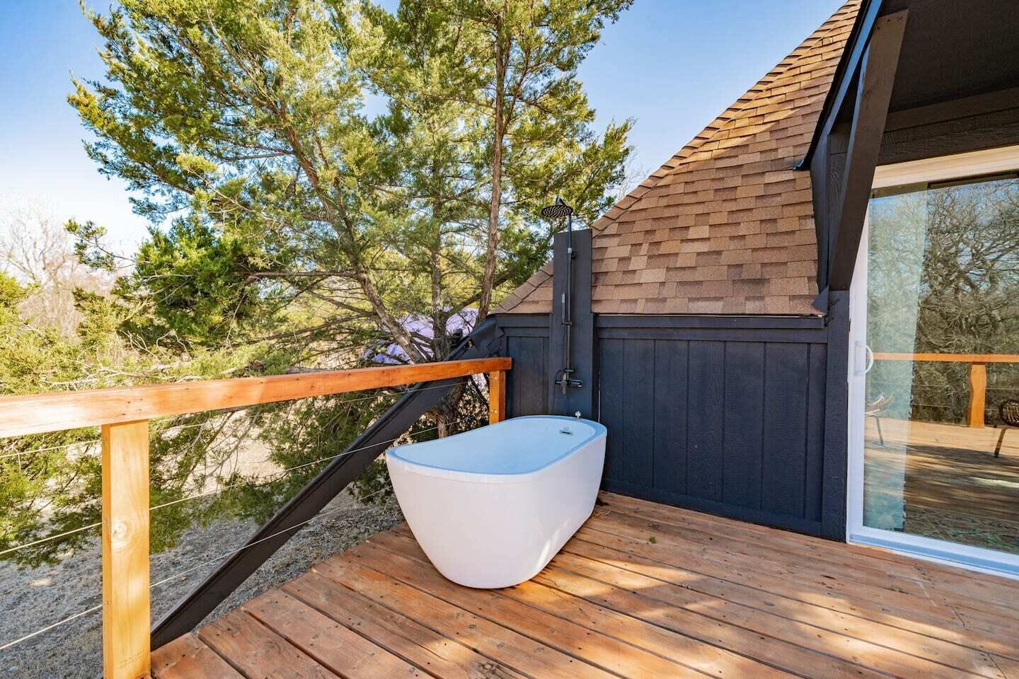 Eine Holzterrasse mit einer weißen Badewanne, einer dunkelholzverkleideten Wand und einer Glastür zu einem Außenbereich.