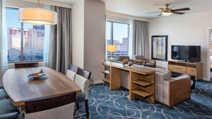 Quarto de hotel com mesa de jantar, cadeiras, sofá, televisão e vista para a cidade.