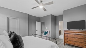 A modern bedroom with a bed, a dresser, a television, and a ceiling fan. - Design-Forward 3BR: King Suite, Pet-Friendly Fenced Yard, Office, Near Greenway (Raleigh)