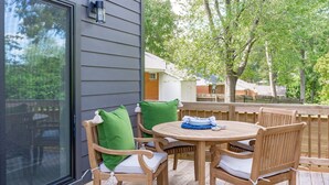 A wooden deck with a round table and chairs, a glass door, and a view of trees and houses. - Design-Forward 3BR: King Suite, Pet-Friendly Fenced Yard, Office, Near Greenway (Raleigh)