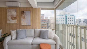 Una sala moderna con un sofá, dos almohadas, un pequeño banquillo naranja y un amplio ventanal con vistas a un paisaje urbano.