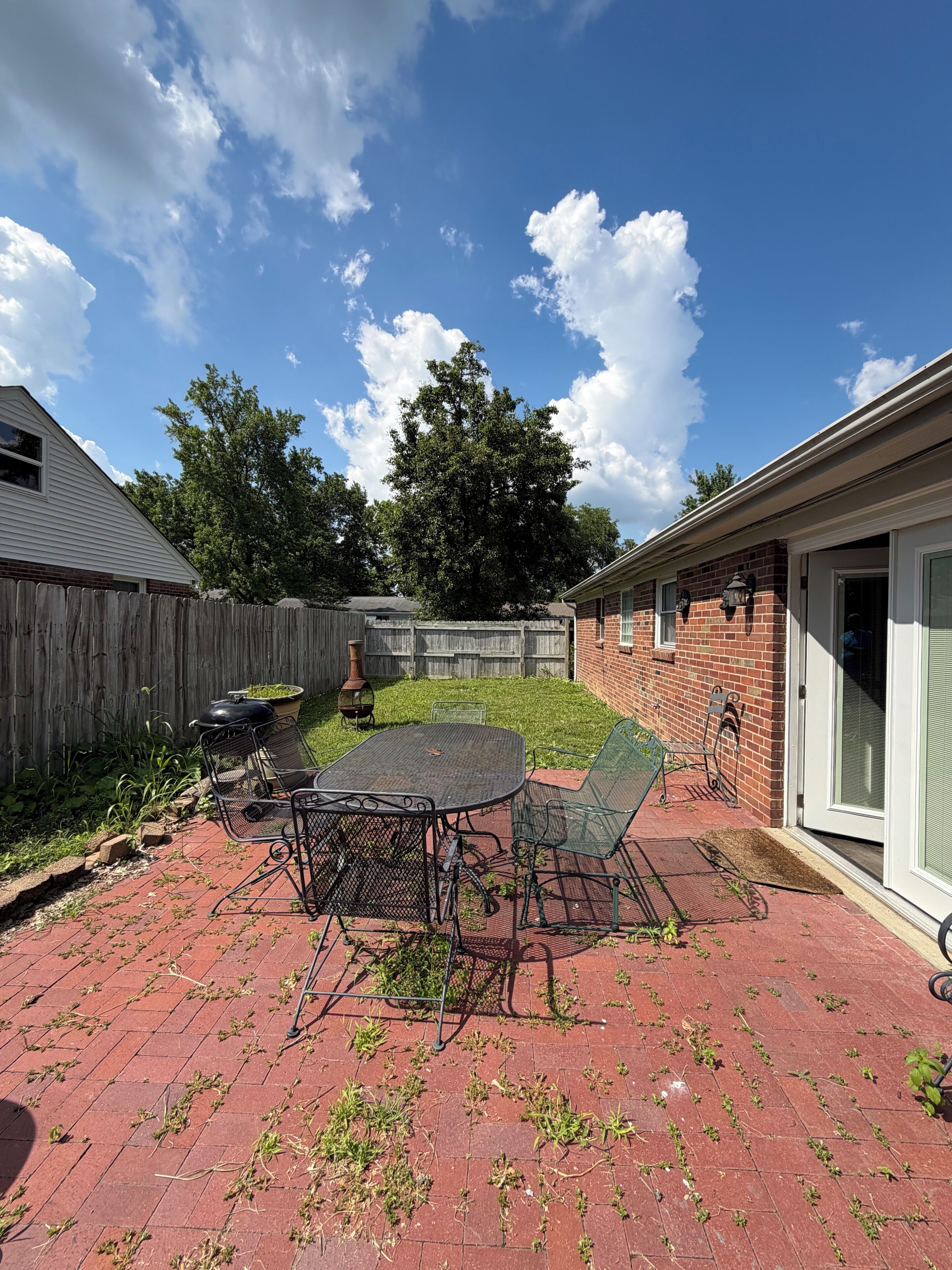 Une terrasse à l’arrière de la maison, avec un sol en briques, une table et des chaises en métal, une clôture en bois et un bâtiment en briques doté d’une porte.