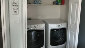 A laundry room with two front-loading washing machines, a shelf above with cleaning supplies, and a sign on the wall reading "Good Food Good Friends Good Times". - PERFECT MIAMI STAY 4-bedroom 2 bath with luxury shower in O (Oxford)