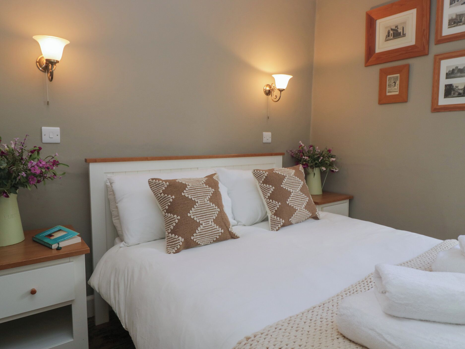 A neatly made bed with two patterned pillows, a bedside table with a book and a vase of flowers, and wall-mounted lamps.