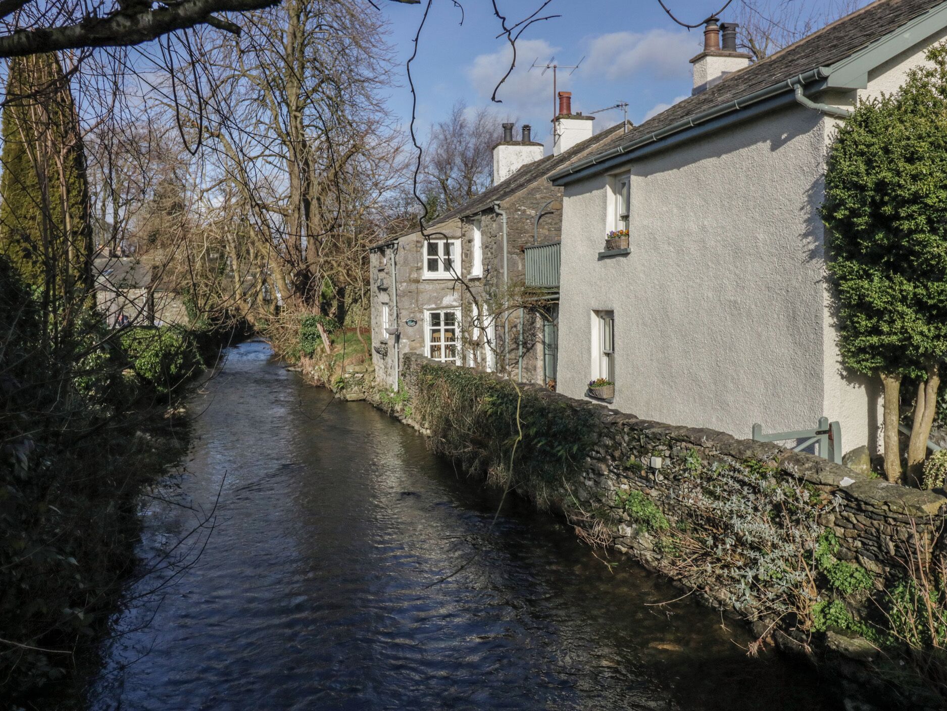 Een smalle waterweg begrensd door stenen muren en traditionele gebouwen met schoorstenen, wat een historische of landelijke omgeving doet vermoeden.